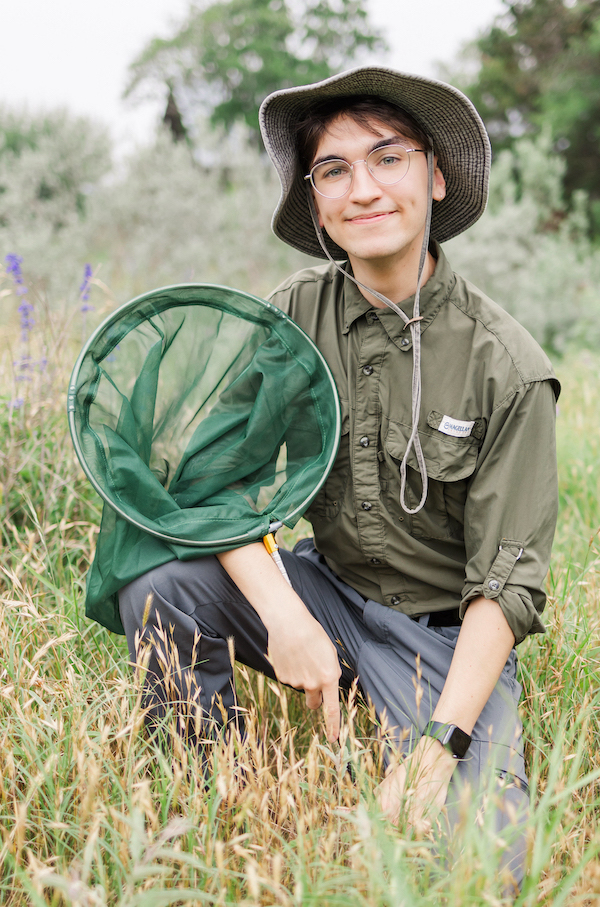 Young man wearing field clothes and hat, kneeling in a field and holding an insect net