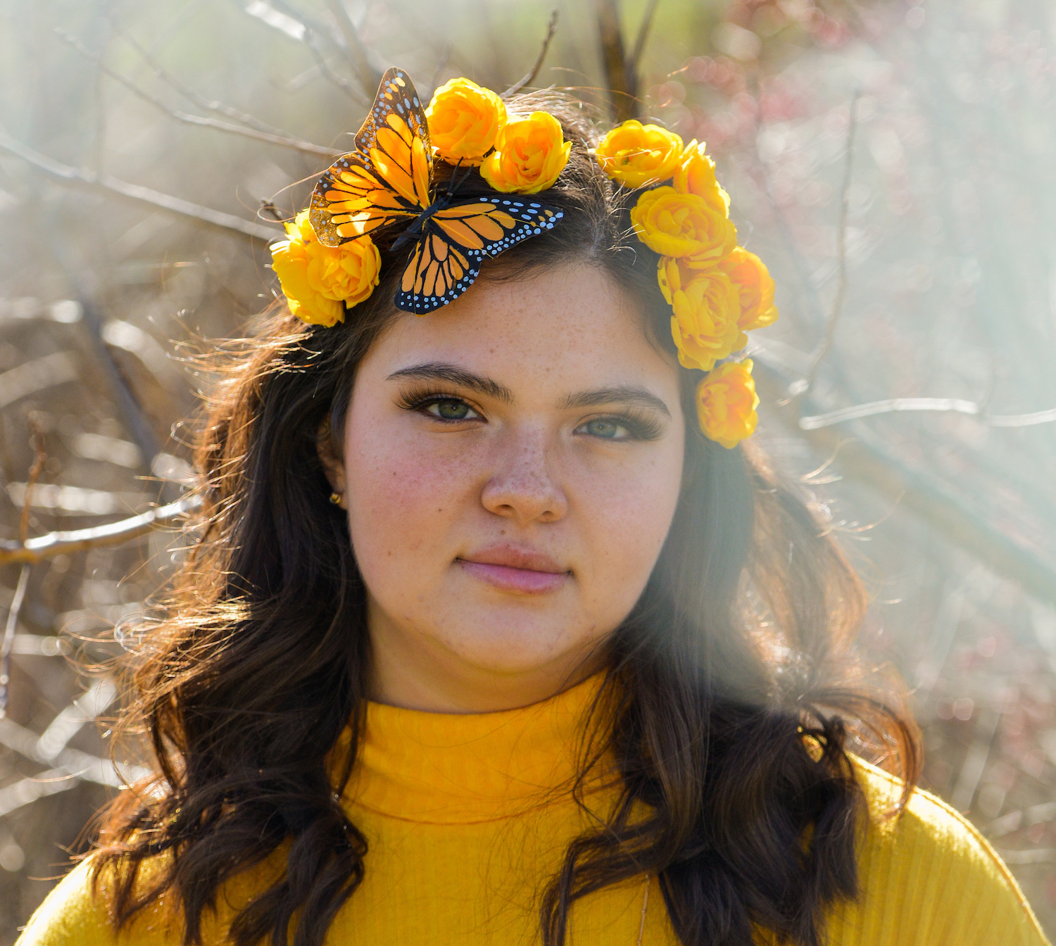 Young woman with dark hair wearing a yellow sweater. She has yellow flowers and a fabric monarch butterlfy in her hair.